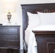 A bedroom scene with a wooden bed, white bedding, and a matching nightstand. The nightstand features three drawers and holds a lamp, phone, and clock.