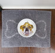 A ceramic bowl with a bulldog face design is placed on a gray textured mat, which lies on a wooden floor.