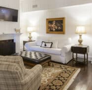 A cozy living room with white wood-paneled walls, a white sofa, beige armchair, ottoman, fireplace, and wall-mounted TV. A framed painting and two lamps on side tables complete the decor.