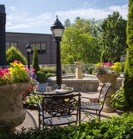 A garden scene with a black wrought-iron table and chairs, surrounded by large flower pots and greenery, featuring a lamp post and a stone fountain in the background.