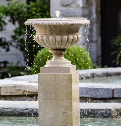 Stone fountain with water flowing over the edges, set in a garden with greenery and stone walls in the background.