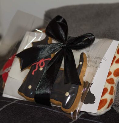 Gift with a black bow containing a cookie shaped like a dress, a card, and an item with orange spots, placed on a gray surface.