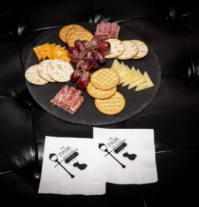 A platter with crackers, cheese, grapes, and sliced sausage on a black surface, accompanied by two branded napkins.