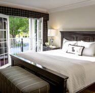 Bright bedroom with a large bed, white linens, and dark wood furniture. French doors open to a balcony with a chair overlooking a lush green view.