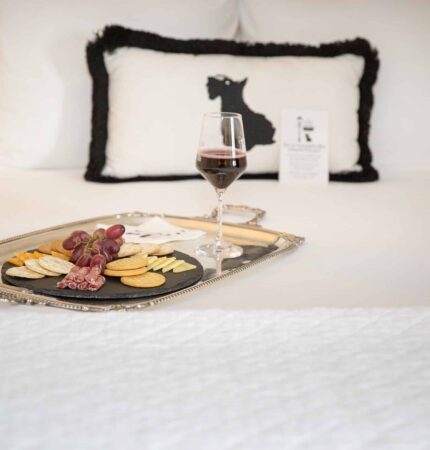 A tray with crackers, grapes, and salami is on a bed, accompanied by a glass of red wine. A pillow with a black silhouette design is in the background.