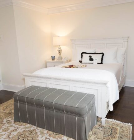 A cozy bedroom with a white bed, patterned pillows, a small nightstand with a lamp, a grey upholstered bench, and a decorative rug on dark wooden floors.