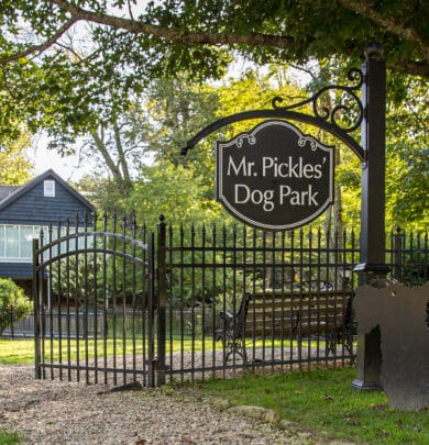 The black wrought iron gate entrance to "Mr. Pickles' Dog Park," adorned with a decorative sign, welcomes visitors amidst trees and a bench, making it one of the charming spots near places to stay in Highlands, North Carolina.