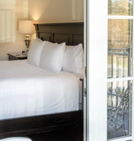 View through a glass door of a neatly made bed with white linens in a well-lit room, with a lamp on the bedside table.