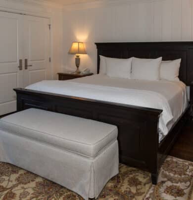 A bedroom with a dark wood bed, white bedding, two bedside tables with lamps, a bench at the foot of the bed, and a patterned rug on dark wood flooring.