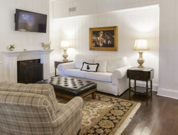 Cozy living room with a white sofa, plaid armchair, and dark leather ottoman. A painting and a wall-mounted TV are above the sofa. Two lamps and a fireplace add warmth.