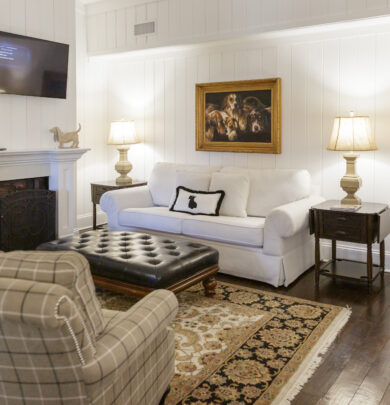 Cozy living room with a white sofa, plaid armchair, and dark leather ottoman. A painting and a wall-mounted TV are above the sofa. Two lamps and a fireplace add warmth.