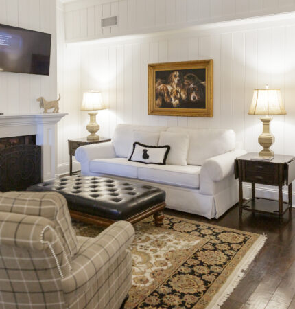 Cozy living room with a white sofa, plaid armchair, and dark leather ottoman. A painting and a wall-mounted TV are above the sofa. Two lamps and a fireplace add warmth.