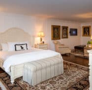 Elegant bedroom with a large bed, cushioned bench, wooden furniture, decorative lamps, and framed artwork on white paneled walls. A floral rug covers the dark wood floor.