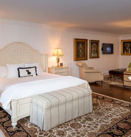 Elegant bedroom with a large bed, cushioned bench, wooden furniture, decorative lamps, and framed artwork on white paneled walls. A floral rug covers the dark wood floor.