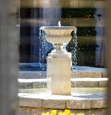 Stone fountain with water flowing, set against a blurred background of a building facade and greenery.