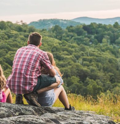 A man and two children sit on a rocky hill, overlooking a scenic view of green forests and distant mountains.