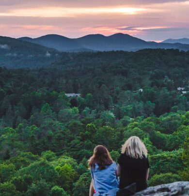 Two people sit facing a scenic view of a forested landscape with mountains in the distance at sunset.