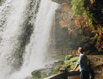 A person stands on a path beside a large, flowing waterfall surrounded by greenery and rocky cliffs.