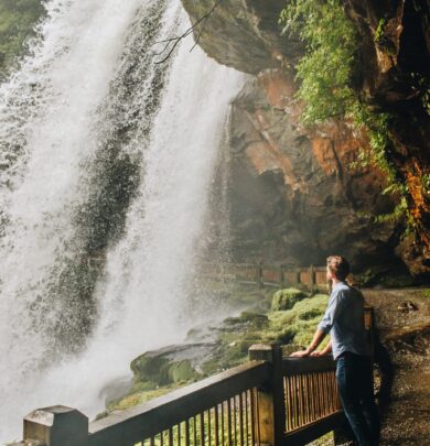 A person stands on a path beside a large, flowing waterfall surrounded by greenery and rocky cliffs.