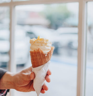 A hand holds a waffle cone with soft serve ice cream and toppings, in front of a window with a blurred view of cars outside.