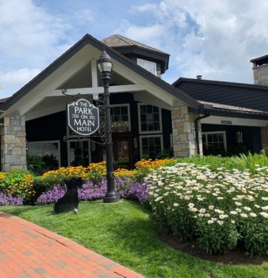 The image shows the exterior of The Park on Main Hotel, featuring stone architecture, a sign, and a well-maintained garden with colorful flowers and a brick pathway in front.