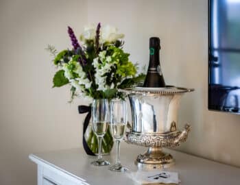 Silver ice bucket with champagne, two glasses, and a bouquet of white and purple flowers on a white dresser.