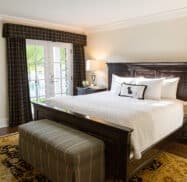 A cozy bedroom with a dark wood bed, white bedding, an upholstered bench at the foot, two nightstands with lamps, a window with plaid curtains, and a patterned rug on the floor.