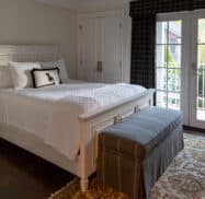 A cozy bedroom with a white bed, matching bedside table, and a dark bench at the foot. French doors open to a balcony with patio furniture. Dark wood floor and patterned rug complete the scene.