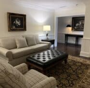 A living room with a beige sofa, plaid armchair, and black tufted ottoman on a patterned rug, featuring paintings of dogs on the walls and a side table with a lamp.