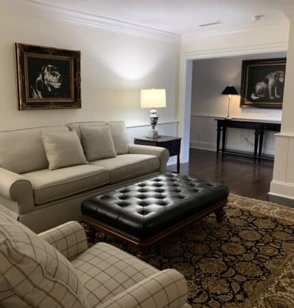 A living room with a beige sofa, plaid armchair, and black tufted ottoman on a patterned rug, featuring paintings of dogs on the walls and a side table with a lamp.